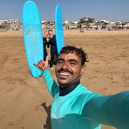Momo and two girls smiling holding their surf boards at the beach of Sidi Kaouki after finishing a surf lesson