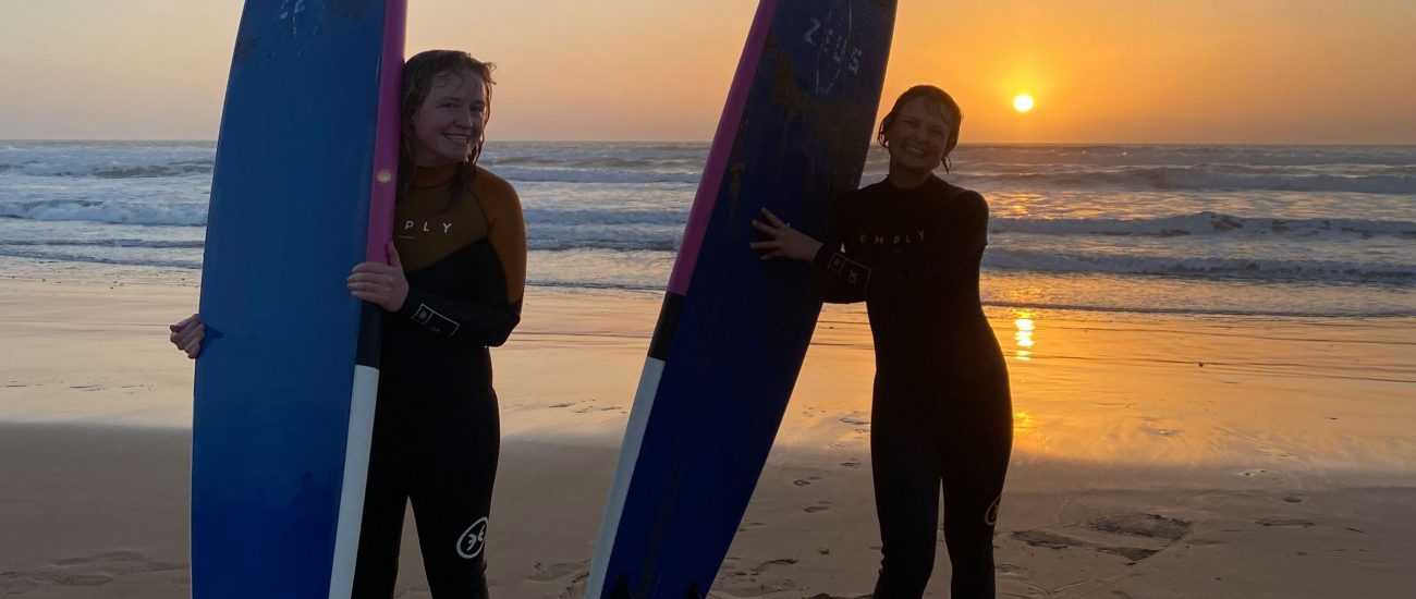 Two girls standing on the beach of Sidi Kaouki during sunset with their surf boards smiling